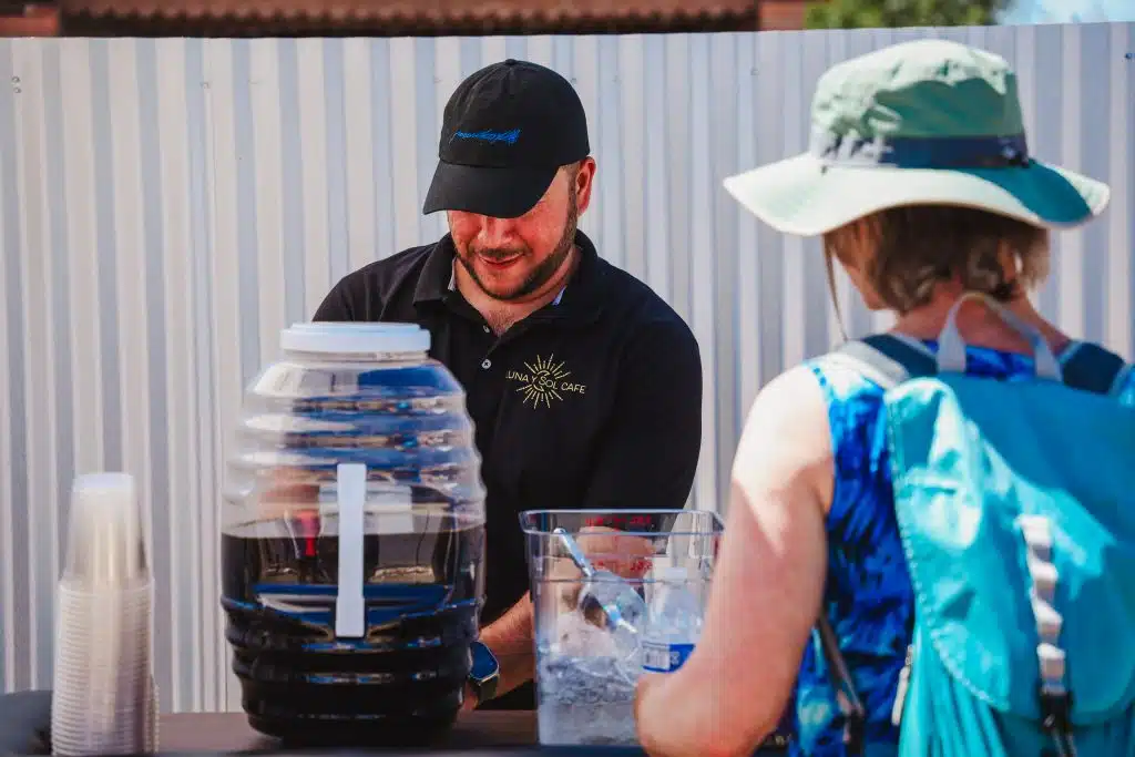 A Luna y Sol Café team member serves drinks at a refreshment station during the community event, with a clear dispenser, ice, and water bottles visible on the table