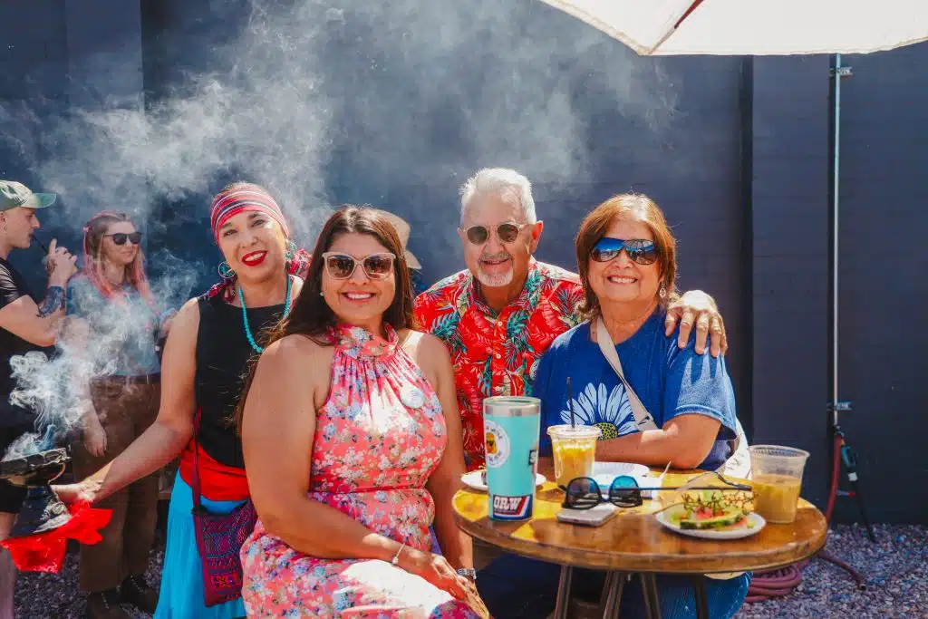 A group of smiling attendees enjoy food and drinks at a table during the Luna y Sol Community Habitat event, with ceremonial smoke rising in the background