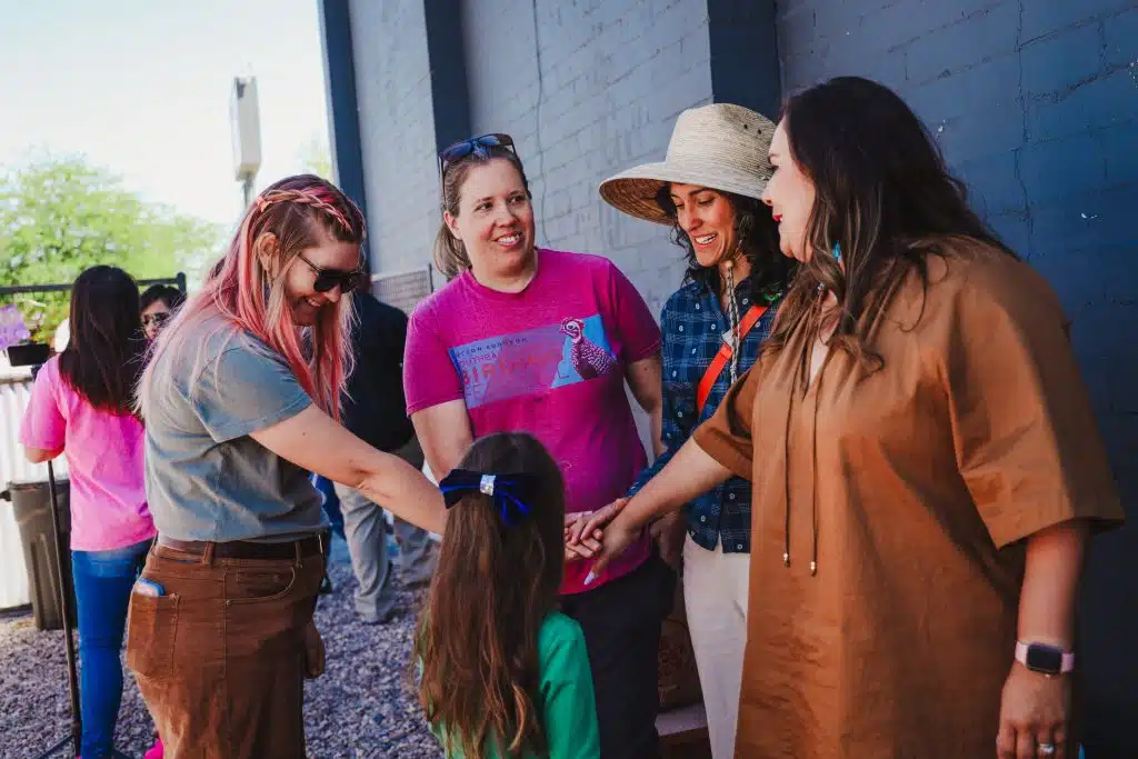A group of women and a young girl join hands in a circle of encouragement and unity during the Luna y Sol Community Habitat event, smiling and gathered against a mural lined wall