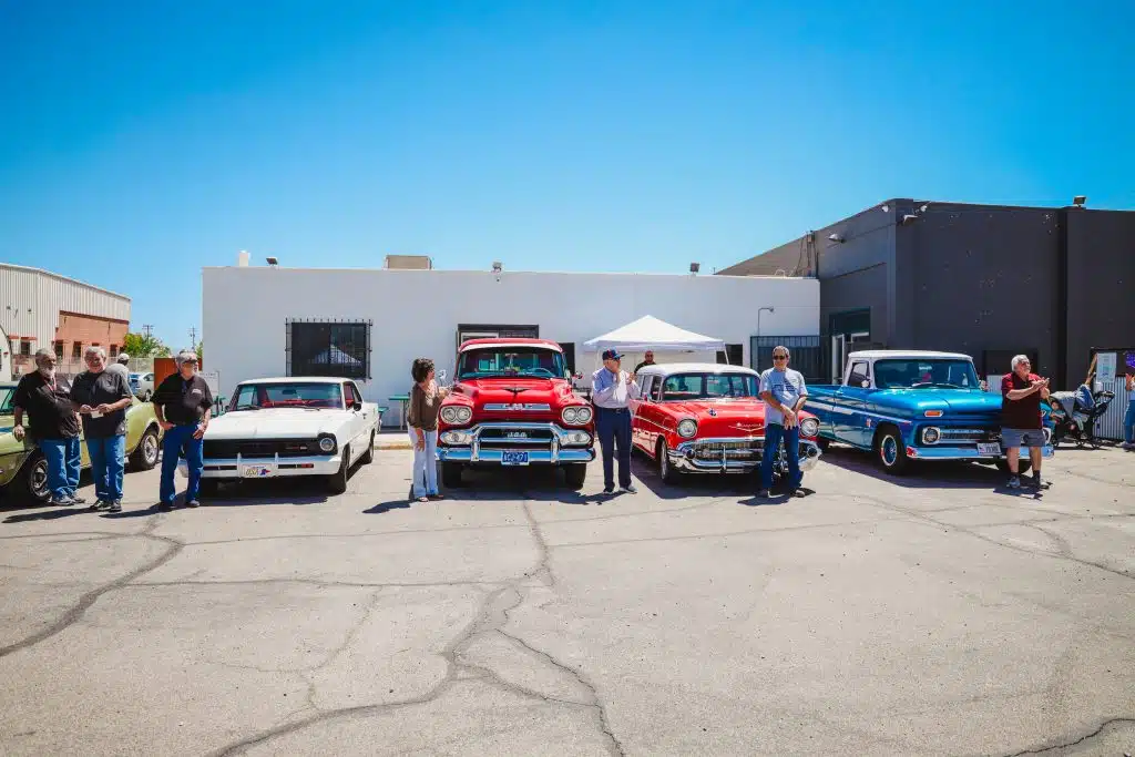 A row of classic cars and trucks displayed at the Luna y Sol Community Habitat event, with owners standing proudly beside their restored vehicles under a clear blue sky