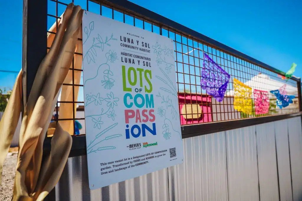 A sign reading “LOTS OF COMPASSION” hangs on the fence at Luna y Sol Community Habitat, with colorful papel picado banners and a bright blue sky in the background