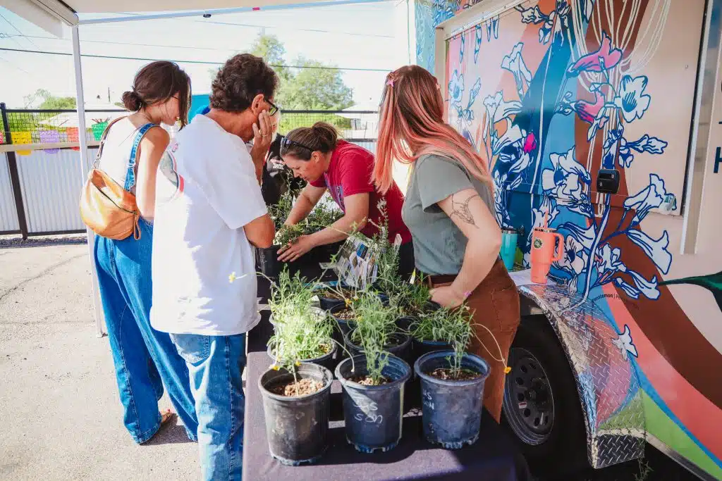 Community members gather around a table of potted native plants at the Luna y Sol Community Habitat event, exploring herbs and greenery in front of a vibrantly painted trailer