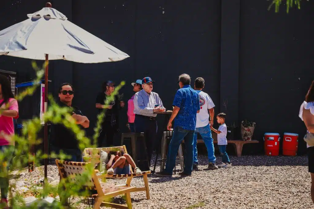 Community members of all ages gather and converse under the shade of umbrellas at the Luna y Sol Community Habitat, with coolers and potted plants