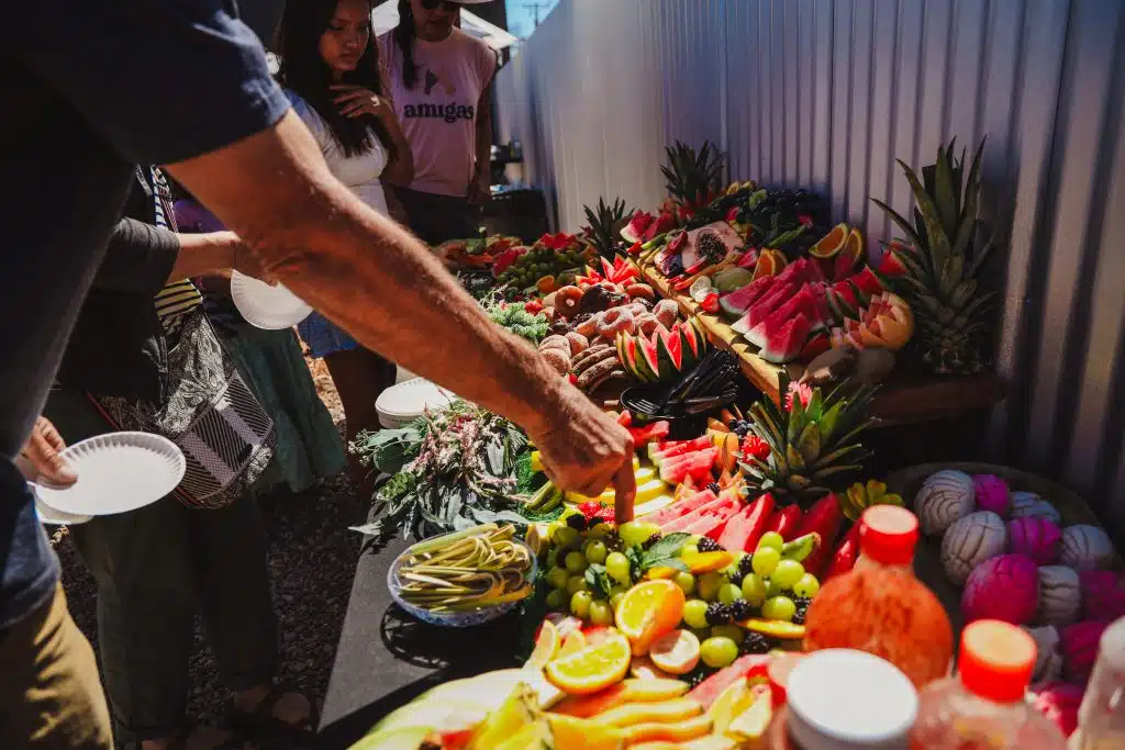 Community members serve themselves from a colorful table filled with fresh fruit, pan dulce, and donuts at the Luna y Sol Community Habitat event, featuring pineapples, watermelon, and vibrant toppings