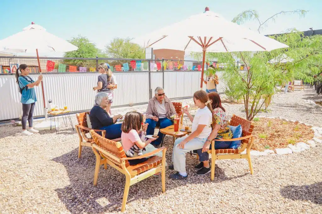 Families and community members enjoy food and conversation under large umbrellas in the garden seating area at Luna y Sol Community Habitat, surrounded by trees, papel picado, and sunlight