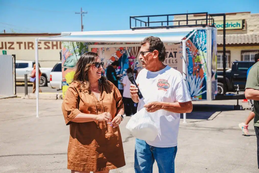 Selina Barajas smiles and talks with a community member holding a microphone and bag, standing in front of the colorful Habitat on Wheels trailer during the Luna y Sol Community Habitat event