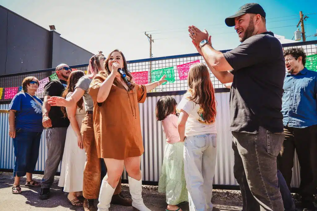 Selina Barajas speaks into a microphone while surrounded by community members and children at the Luna y Sol Community Habitat event, standing in front of colorful papel picado and receiving applause