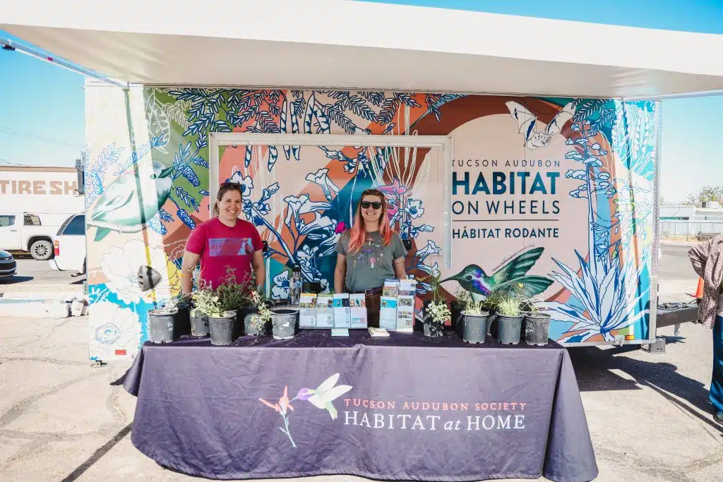 Two women stand behind a Tucson Audubon Society Habitat on Wheels display at the Luna y Sol Community Habitat event, with native plants, brochures, and a vibrantly illustrated trailer