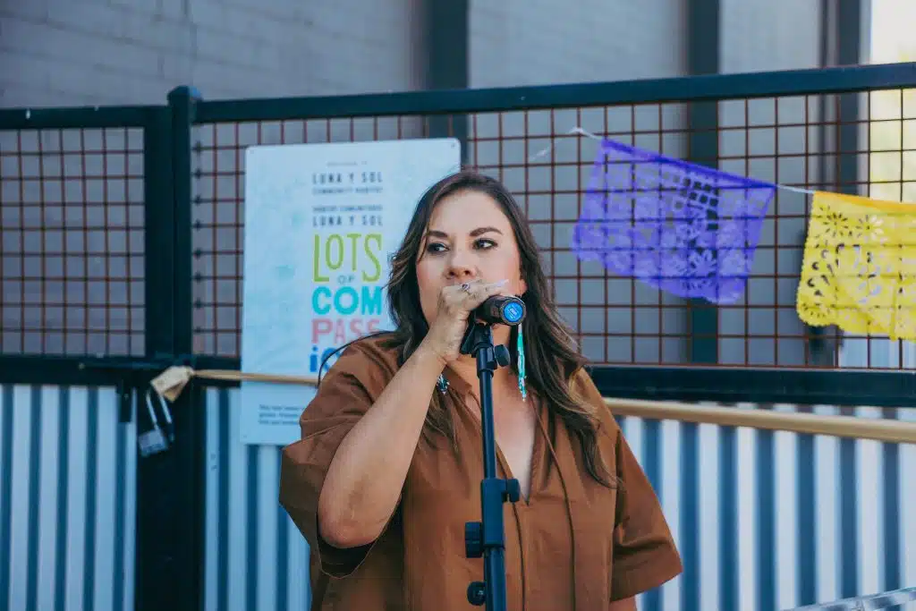 elina Barajas speaks passionately into a microphone during the Luna y Sol Community Habitat event, standing in front of papel picado and a “Lots of Compassion” garden sign