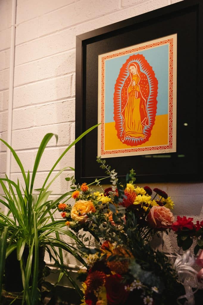 A framed screen print of the Virgen de Guadalupe hangs on a brick wall above fresh flowers and plants.