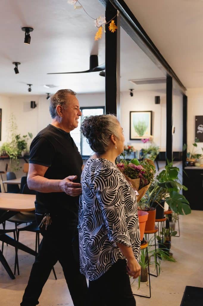 A man and a woman walk through the cafe past tables and potted plants, she carrying a bouquet in brown paper