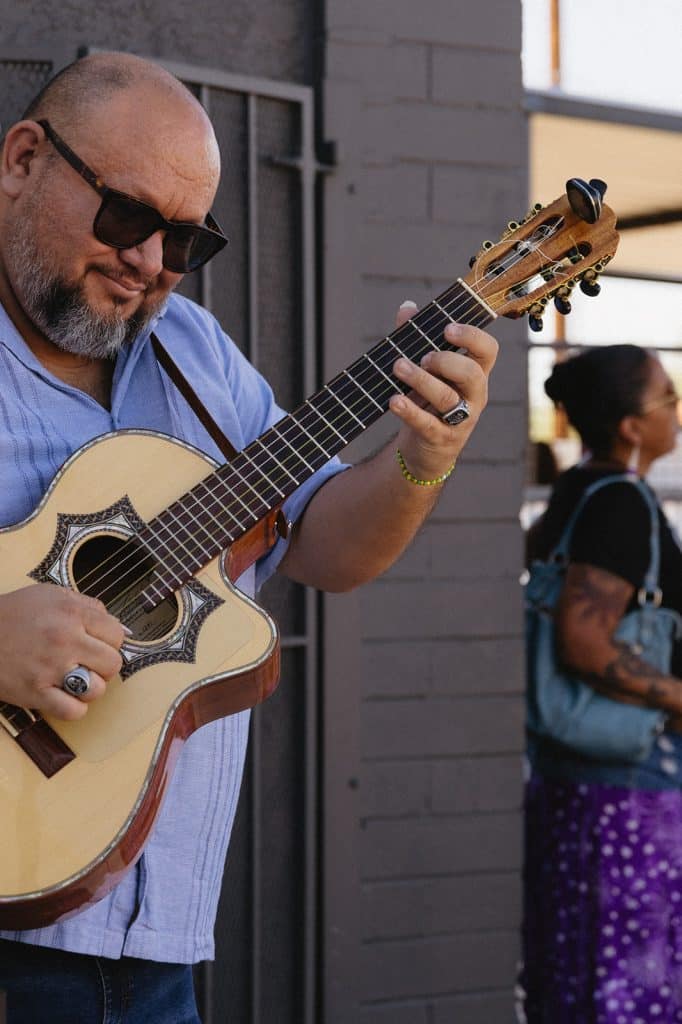A man in sunglasses and a light blue shirt plays an acoustic guitar with an inlaid sound hole, a woman behind him.