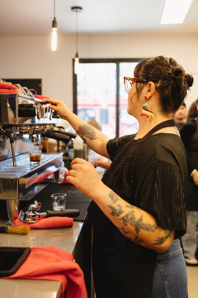 A person in a black apron pulls espresso shots, one arm raised to the machine.