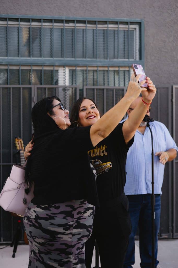 Two women lean in for a selfie beside a microphone stand, with a guitar on a stand and a man in a blue shirt behind.