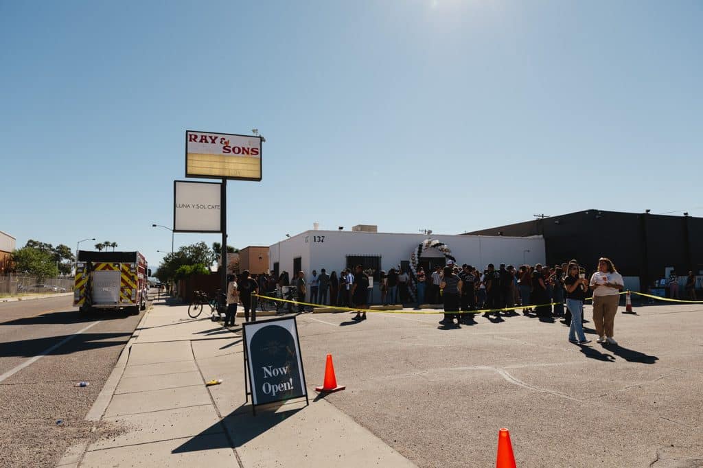 A white corner building with a balloon arch and a crowd out front, a Now Open! sign on the sidewalk, a fire truck at left.