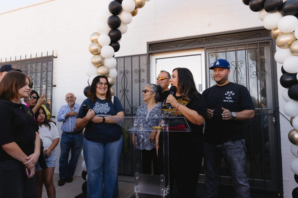 Speakers stand at a clear podium under a balloon arch, with a man in an LA cap and a Luna y Sol, Barrio Libre shirt beside them.