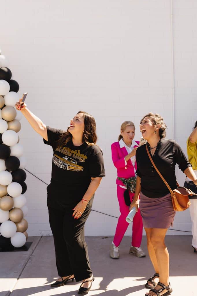A woman in a lowrider shirt holds up her phone and laughs beside a balloon column, another woman laughing next to her.