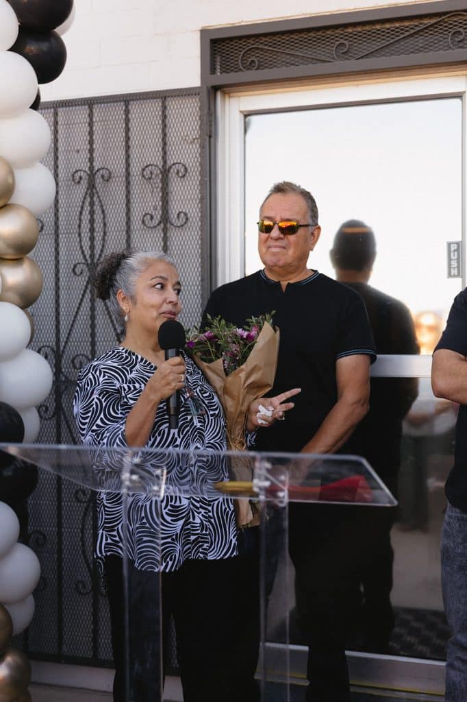A woman in a black-and-white print top speaks into a microphone, bouquet in her arm, as a man in sunglasses looks on.
