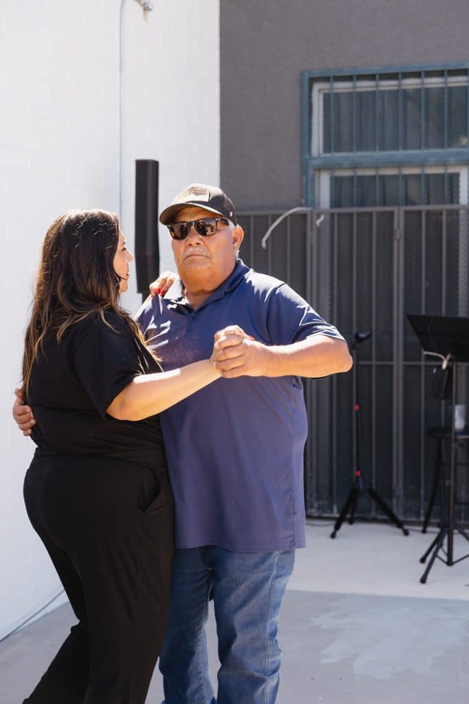 A woman in black and a man in a cap and sunglasses dance close on the patio, a speaker and music stands behind them.