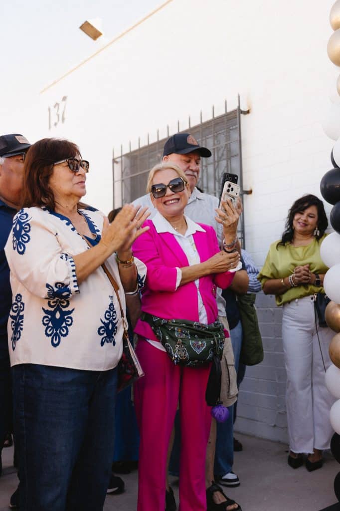 Guests in sunglasses applaud and film on a phone beside a white wall marked 137, with a balloon column at right.