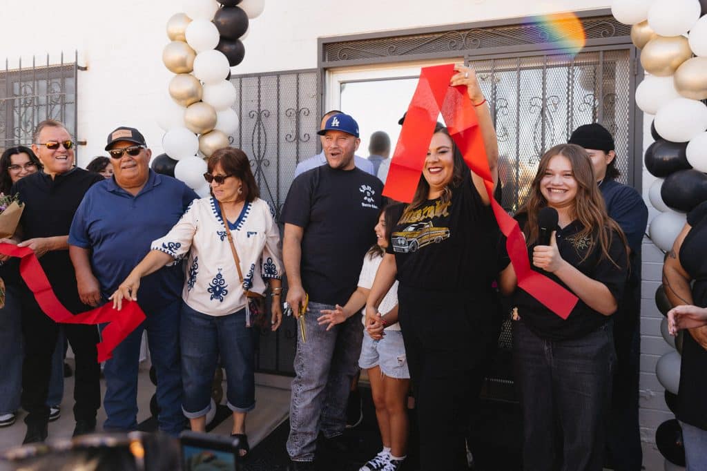 A woman raises a cut red ribbon as people cheer by the balloon arch; a young person holding a mic gives a thumbs up.