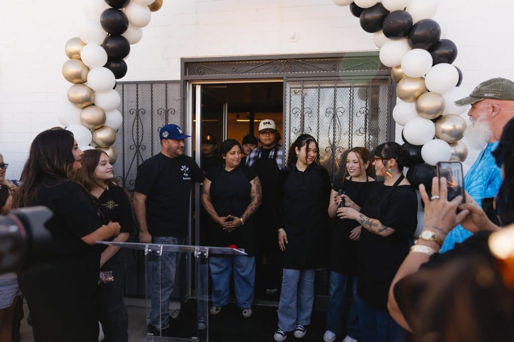 People in black aprons stand in the open cafe doorway under a balloon arch as one speaks into a microphone.
