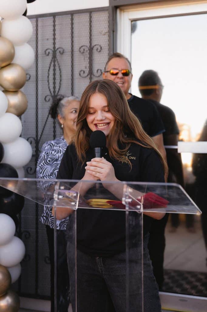 A young person in a Luna y Sol Cafe polo smiles at a microphone, hands clasped, at a clear podium beside balloons.