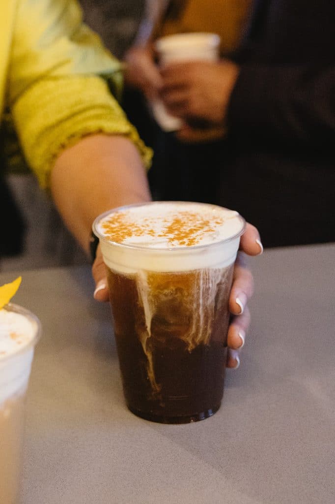 An iced cold brew topped with cream and cinnamon sits on a gray counter as a hand reaches for it.