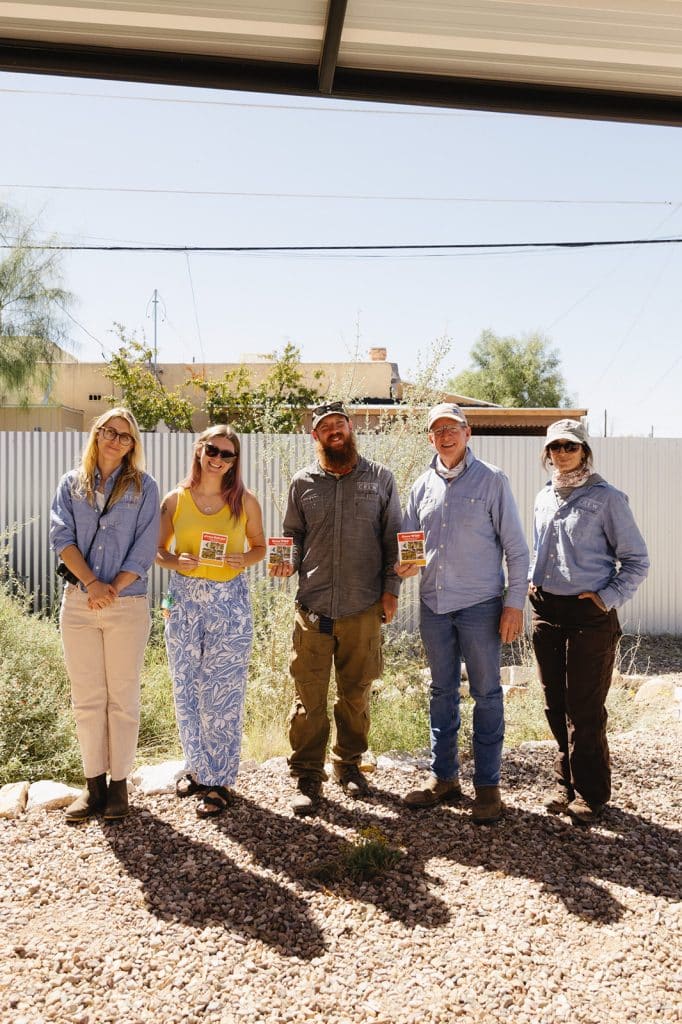 Five people in matching blue work shirts stand in a gravel garden bed holding small booklets, a metal fence behind.