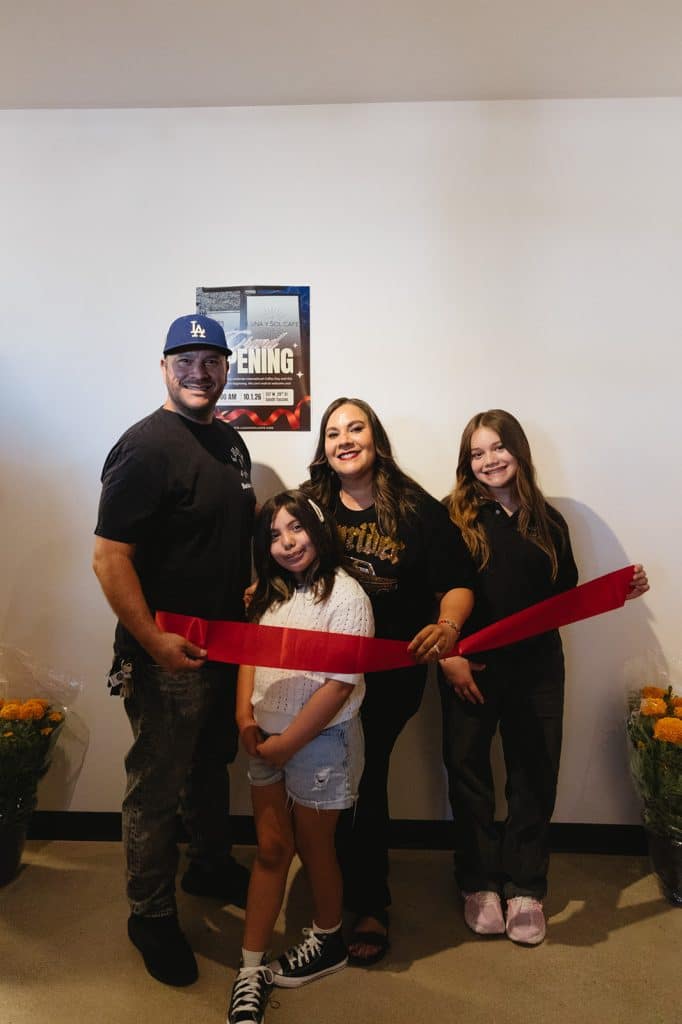Four people hold a red ribbon in front of a Grand Opening poster dated 10.1.26, with orange marigolds on each side.