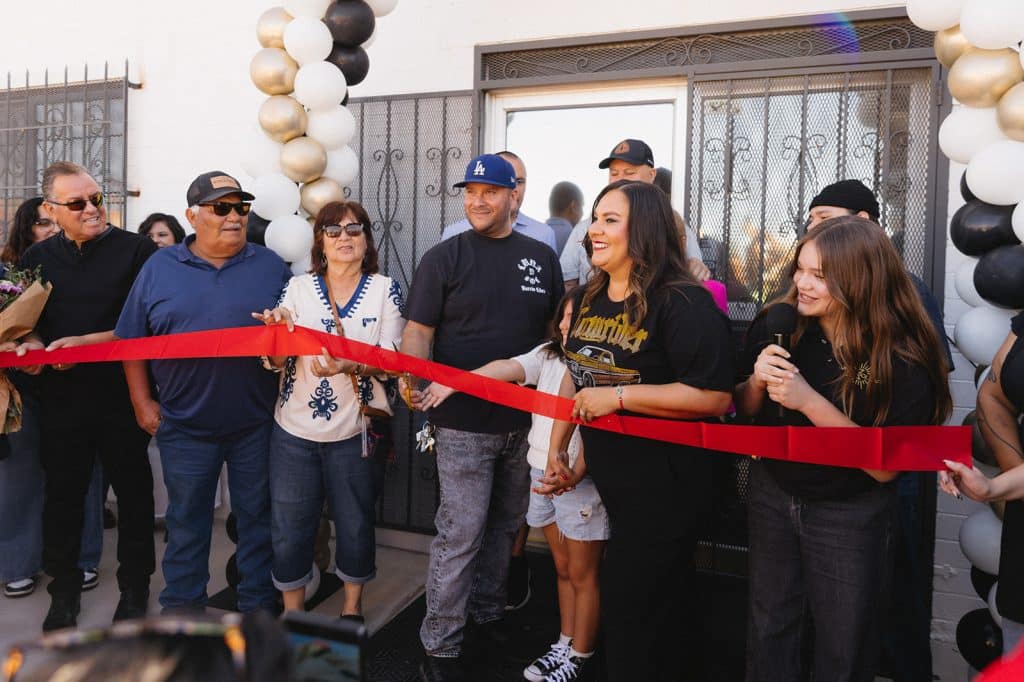 People hold a red ribbon across the doorway under a balloon arch as a young person with a microphone looks on.
