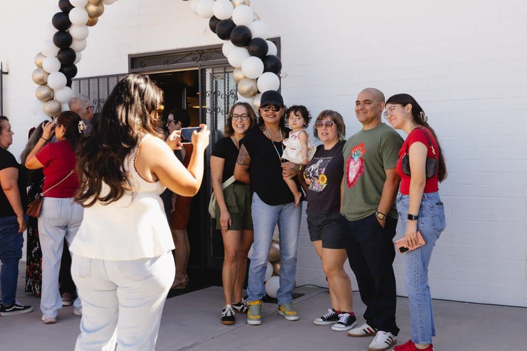 Six people pose by the balloon arch, one holding a toddler, as a woman in white takes their photo with a phone.