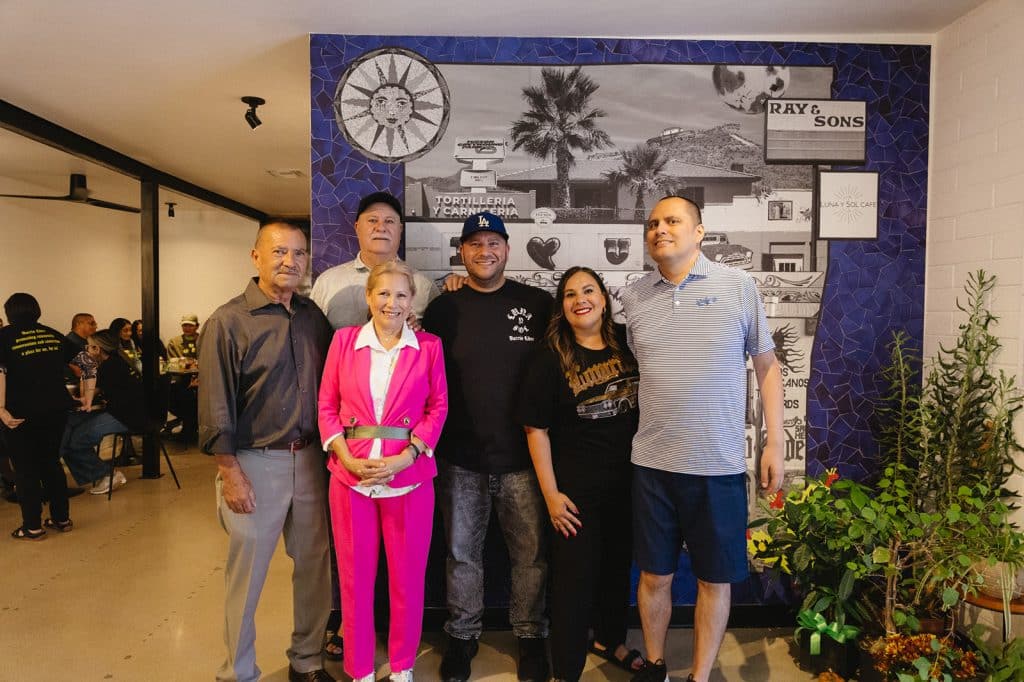Six people pose in front of a blue tile mural of the old Ray & Sons sign, a sun face and a Luna y Sol Cafe sign.