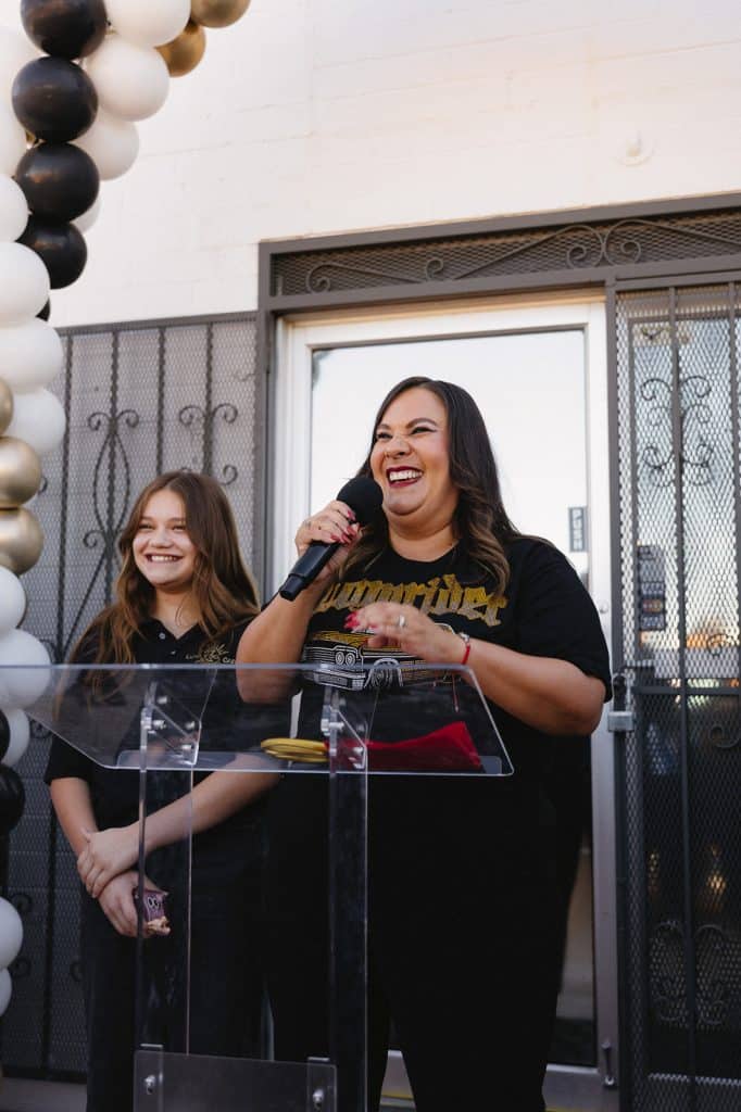 A woman laughs into a microphone at a clear podium with gold scissors and a red ribbon; a young person smiles behind.