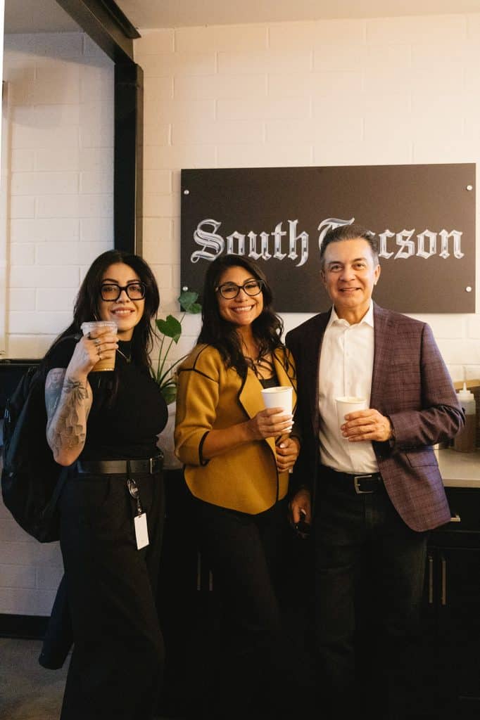 Three people smile holding drinks under a black South Tucson sign on a white brick wall.