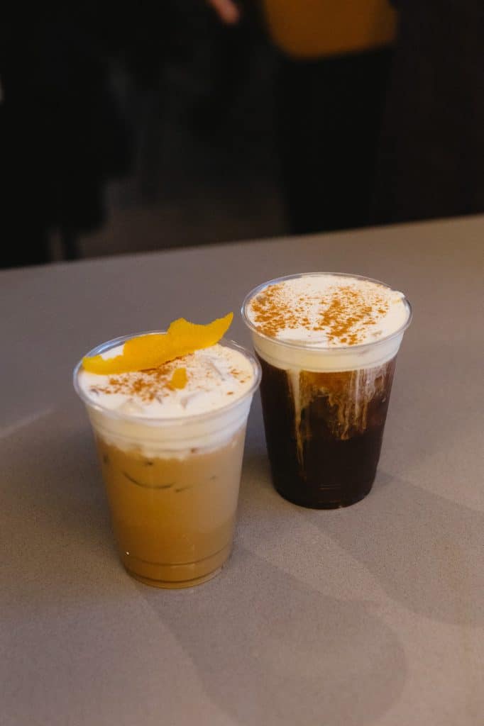 Two iced drinks with cream and cinnamon on a gray counter, one topped with a curl of orange peel.