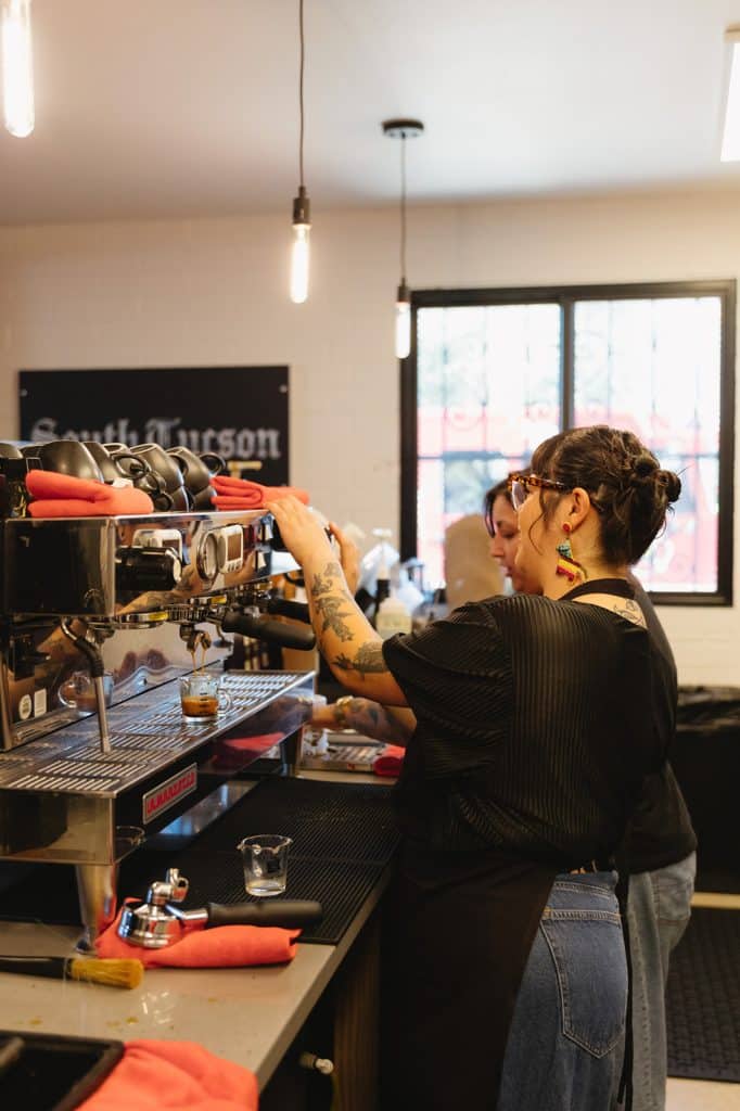 Two people work behind the espresso machine as shots pour into small glasses, with a South Tucson sign on the wall.