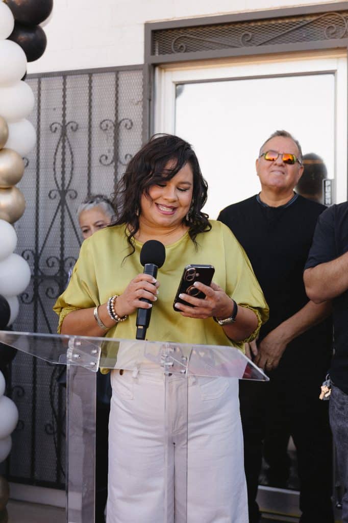 A woman in a yellow-green blouse smiles at her phone while holding a microphone at a clear podium.