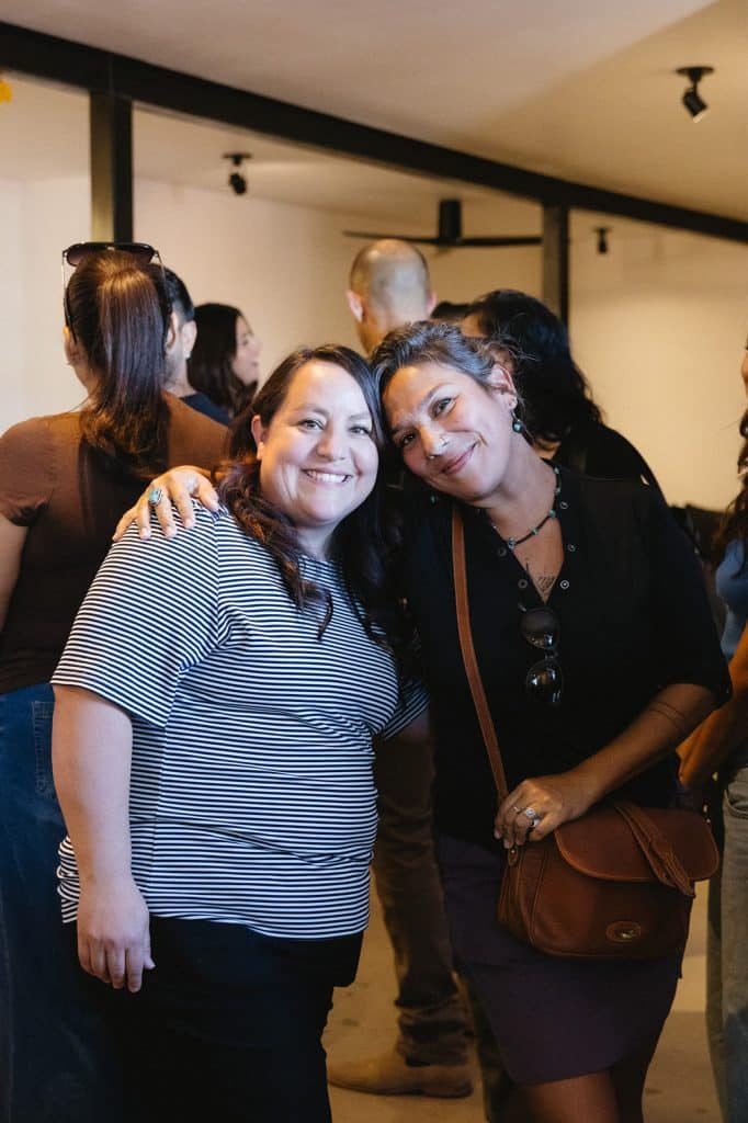 Two women lean their heads together and smile inside the cafe, with guests mingling behind them.