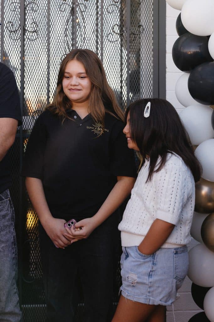 Two young people stand beside a black, white and gold balloon garland, one in a black Luna y Sol Cafe polo.